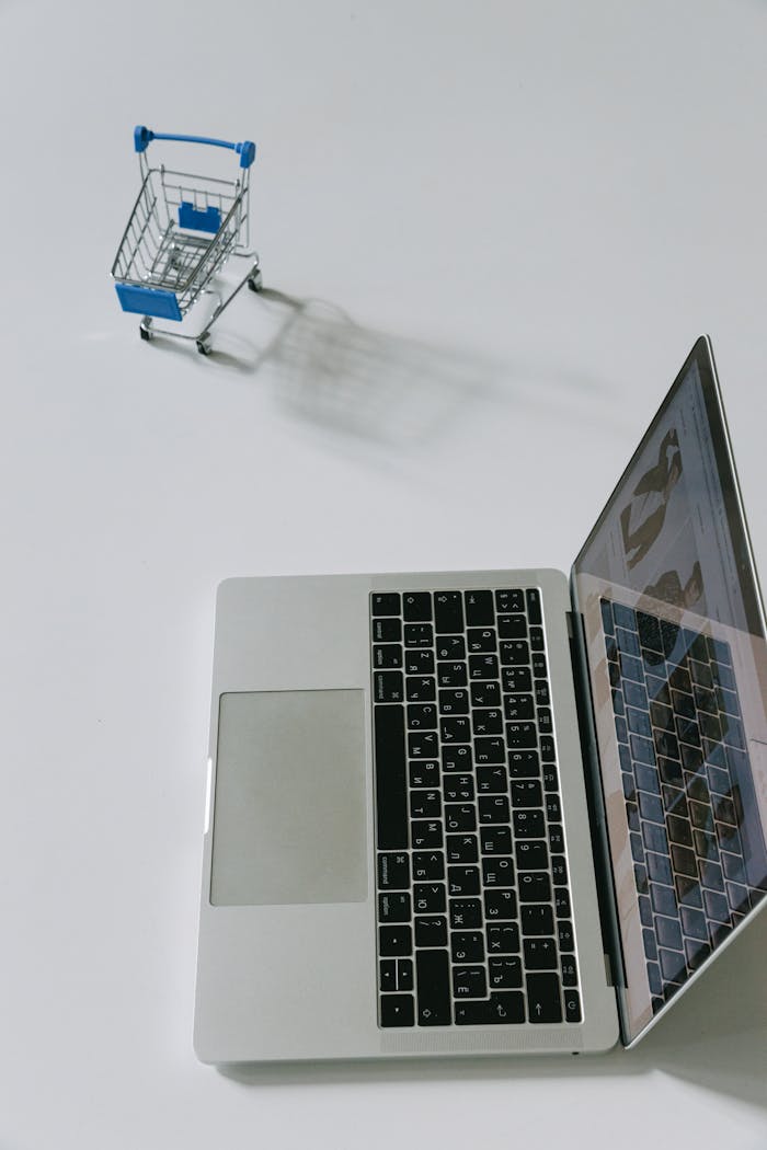 Minimalist scene of a laptop beside a miniature shopping cart symbolizing online shopping.